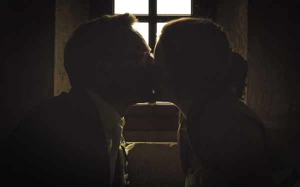 Ein künstlerisches Schwarz-Weiss-Hochzeitsporträt eines Brautpaares als Schattenbild vor einem hell erleuchteten Fenster. Das Paar küsst sich innig, wobei ihre Silhouetten und ein Teil des Fensterrahmens im Kontrast zum Licht stehen. Das Bild vermittelt tiefe Romantik, Intimität und eine stimmungsvolle Atmosphäre der Hochzeitsfotografie von Michael Schalansky in Thüringen.